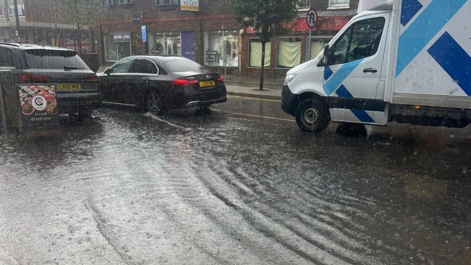 Cars and a van are driving through a flooded street. There are shops in the background