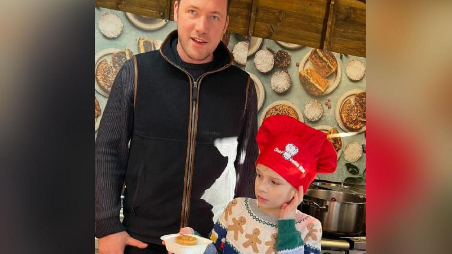 Joshie Harris pictured standing with Tommy Banks in York's Christmas market. He is wearing a red hat and a multi-coloured jumper with gingerbread men on.