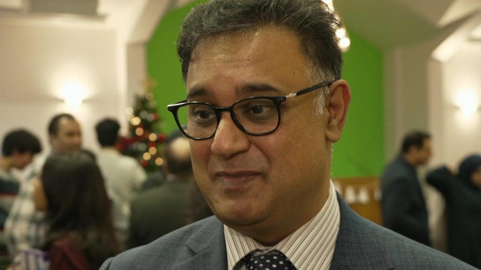 A man with short black and grey hair, who is wearing black glasses and a suit with a striped shirt, stands in a church.