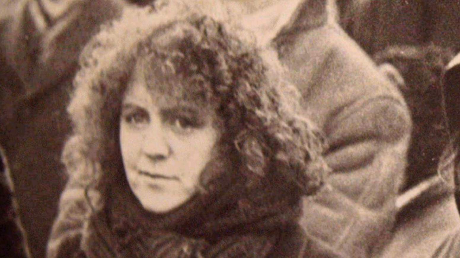 Black and white archive image of Nell McCafferty as a young woman - she has long curly hair and is wearing a scarf wrapped around her neck and chin