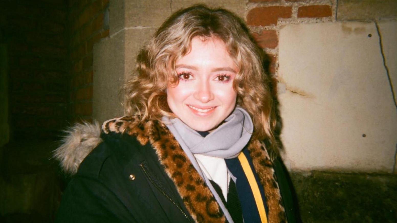 Paloma, pictured in what looks like a flash photo from a film camera, with her face brightly lit smiling in the centre of the frame, her hair in long blond curls, wearing a coat with a furry hood and with something in leopard print faux-fur around her shoulders. She is standing against a brick and stone wall at night
