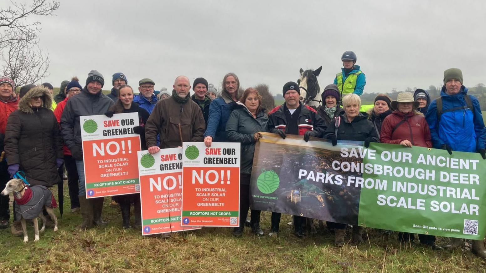 A large group of people in winter coats and hats stand huddled together in a field holding up banners and signs.