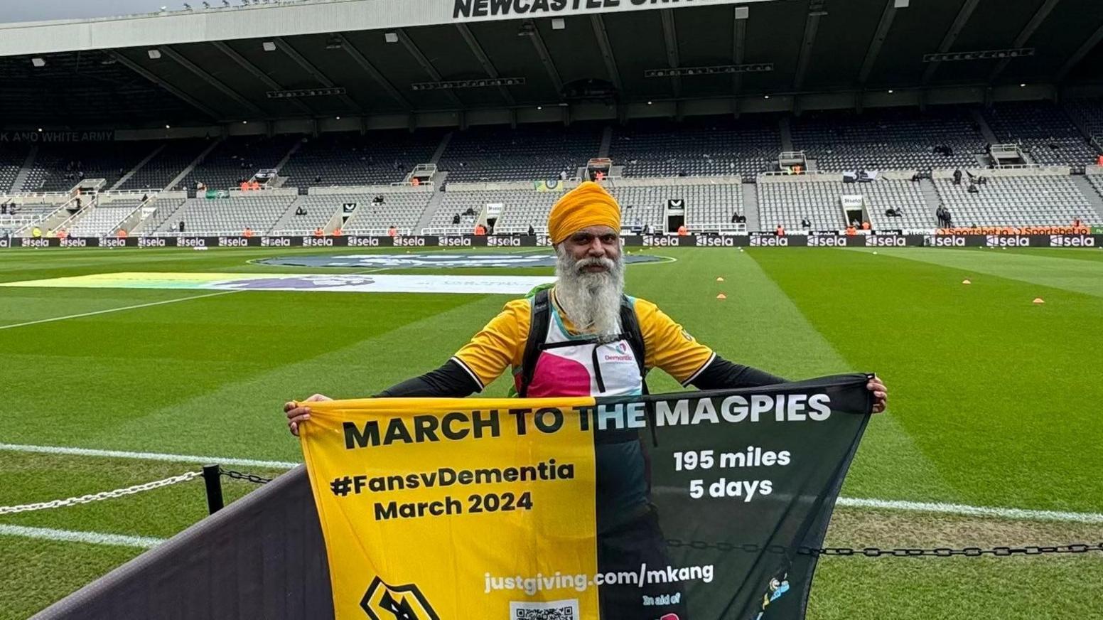 Manny Singh Kang with banner saying "March to the Magpies" on a football pitch inside a stadium. He wears a yellow head covering and has a long white beard