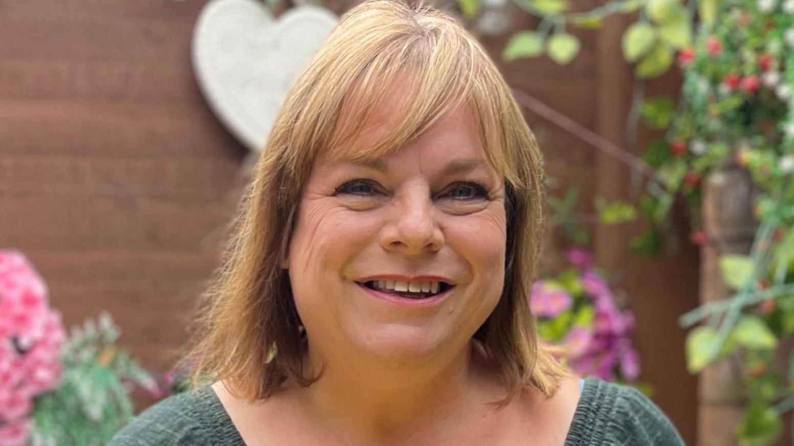 A woman who is standing outside in front of a tree and flowers. She is wearing a green top and is looking directly at the camera and smiling.