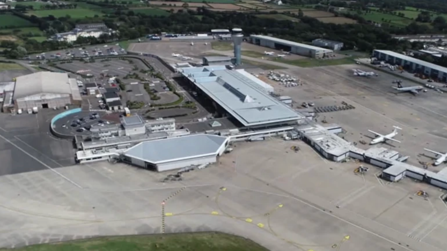 Aerial view of Jersey Airport, a large grey building situated on tarmac. There are a couple of planes waiting at terminals on the right hand side.