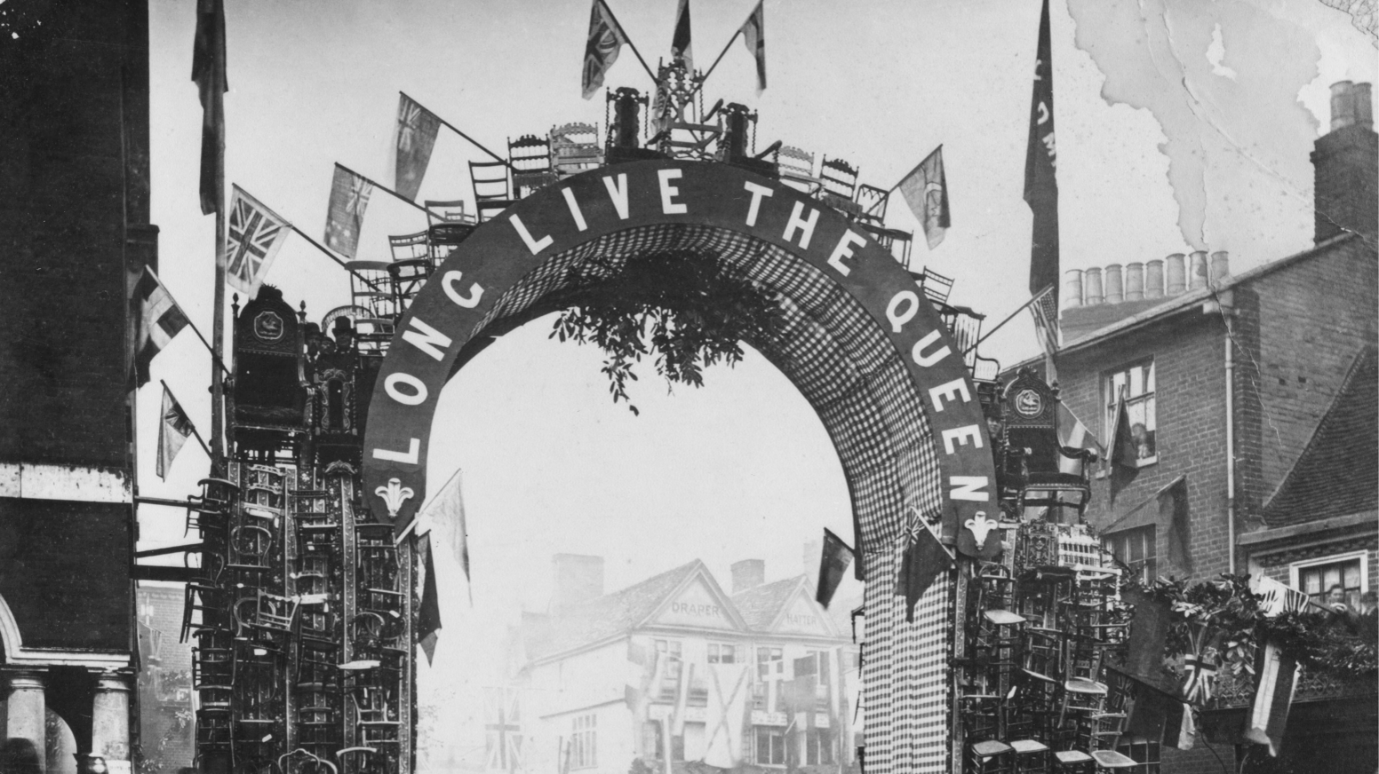 A black and white photo with an arch saying Long Live the Queen, decorated with flags and wooden chairs, in a high street