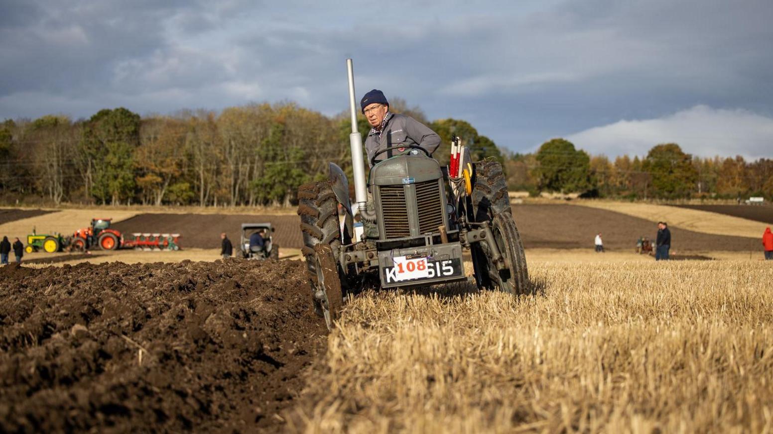 A man drives a vintage tractor as it ploughs a field of stubble. There are other tractors, and also people, in the background the field in lined by trees.