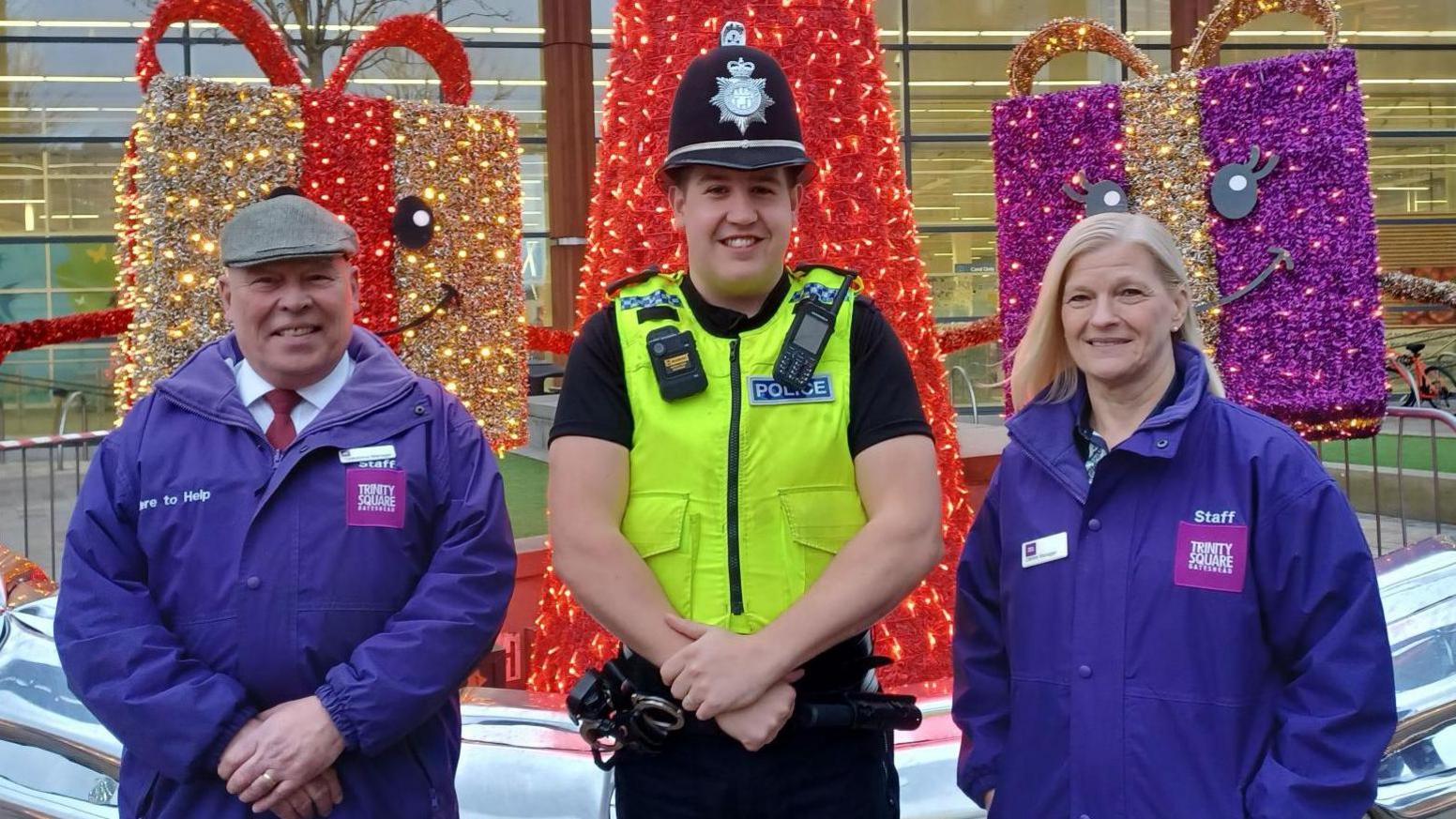 A police officer in full uniform stands next to a man and woman who are wearing purple coats with the logo 'Trinity Square'.