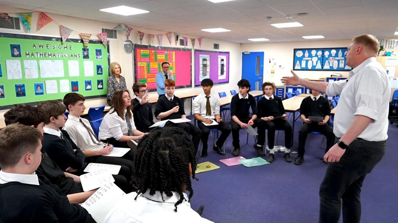 Paul Clark, in white shirt and black trousers, addresses a group of teenage boys in a classroom. The boys are wearing school uniform and their chairs are arranged in a semi-circle.