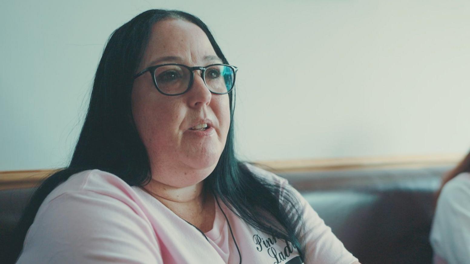 Sarah Jane Waugh, sitting down and wearing a pink tshirt with the Falkirk Pink Ladies group logo. She has long black hair and is wearing glasses.