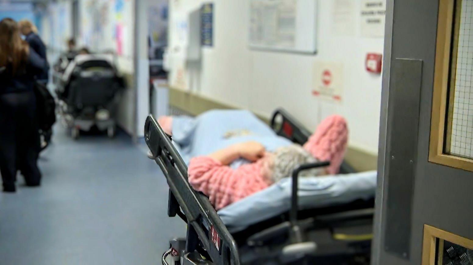 An elderly lady in a pink dressing gown lies on a hospital trolley placed along a corridor. There is another hospital bed with a patient further down the corridor and people are walking past them as they lie there.