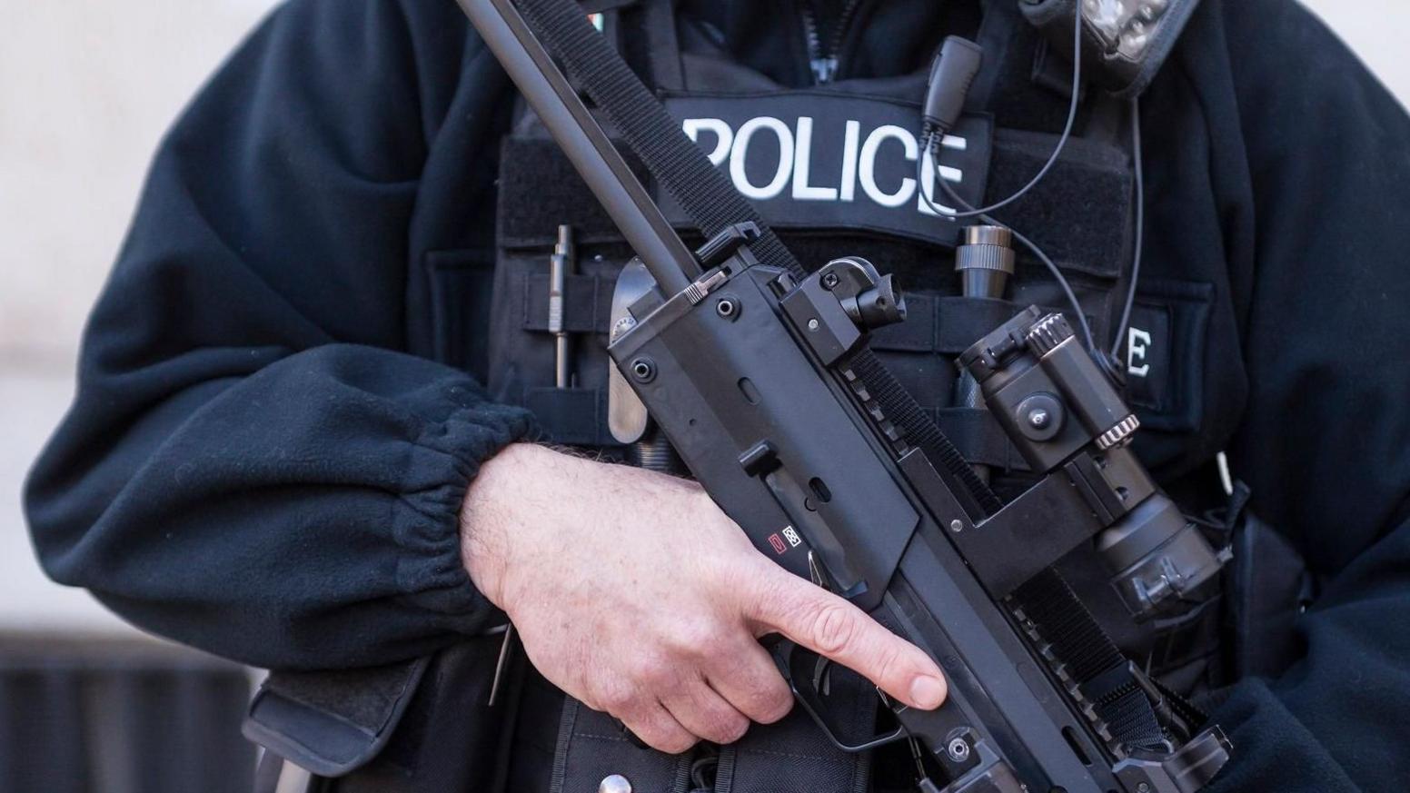 An armed police officer's mid section. He is dressed in black with a bullet-proof police vest on and is holding a large gun