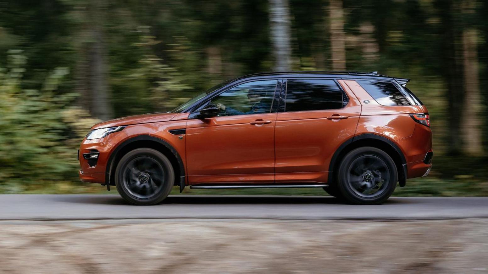 An orange-coloured Land Rover drives along a road with trees blurred beyond it.