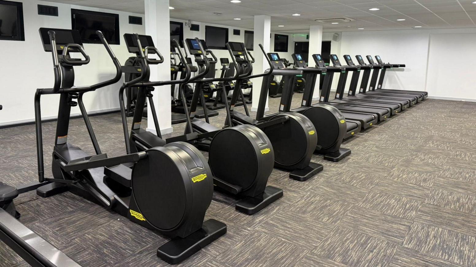 A row of brand new black fitness machines in a gym. To the forefront are four elliptical machines, behind are eight treadmills. There are also various other machines, with large TV screens on the walls.