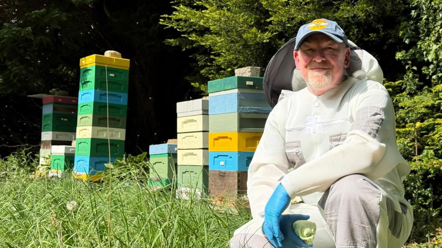 Beekeeper Ashley Galley wears a protective white suit next to series of coloured hives.