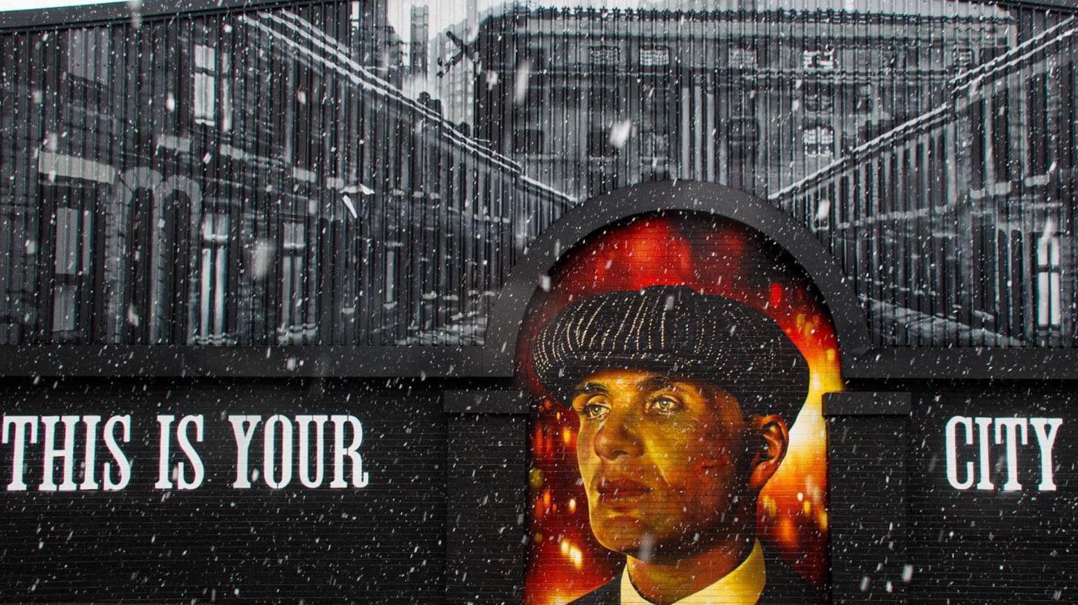 The mural is painted black and says 'this is your city'. The top of the factory wall is filled with painted black and white terraced houses. Thomas Shelby is painted in an archway of the wall.