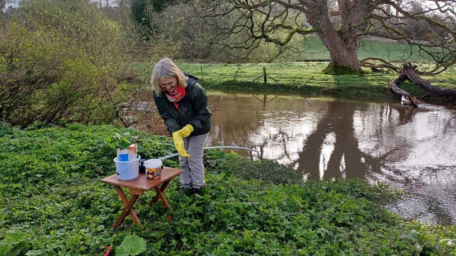 A woman is standing in front of a small foldup table on a riverbank. She is wearing yellow gloves and is about to do some testing 