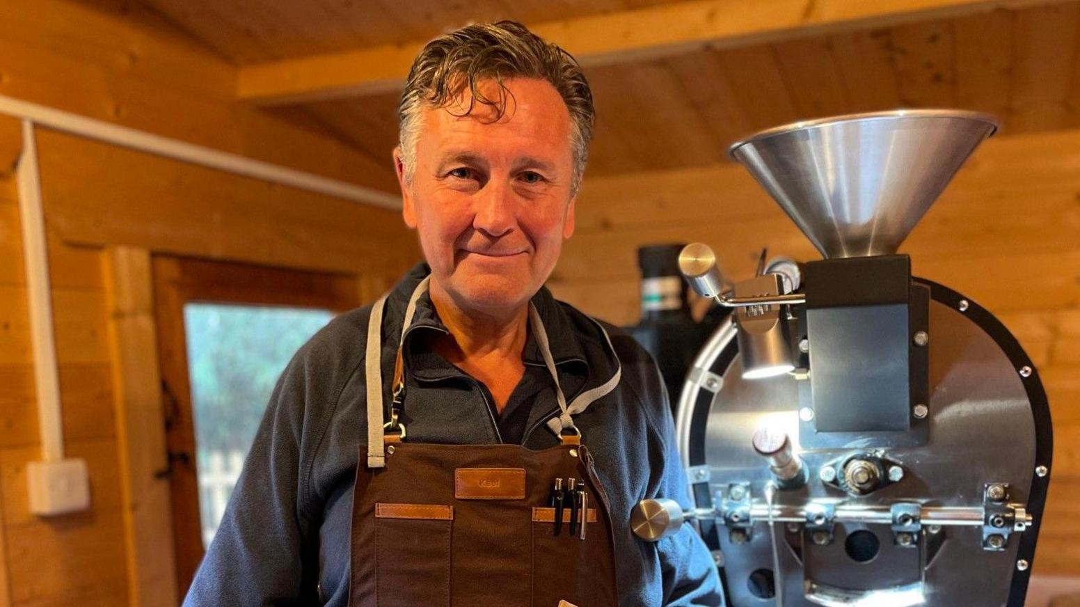 A man with grey hair, wearing a brown apron and blue jumper and smiling at the camera. Standing next to a silver coffee roasting machine. He is in a wooden out building