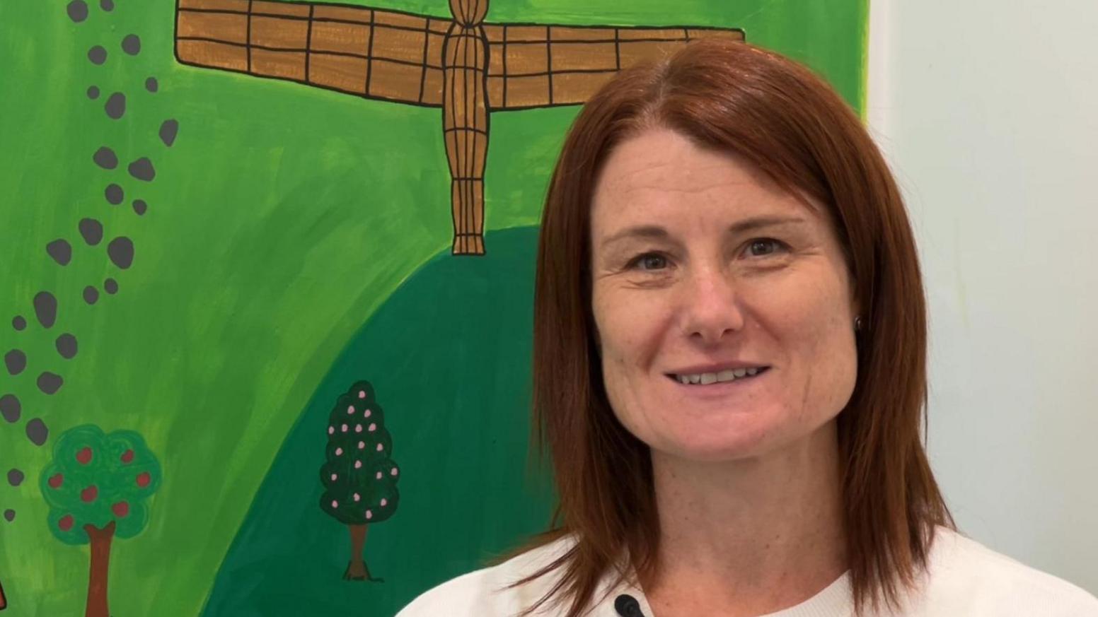 Tracey Guy has shoulder-length reddish brown hair. She is smiling directly into the camera and standing to the side of a childlike drawing of trees and parkland. There is also a drawn image of the Angel of the North just behind her.