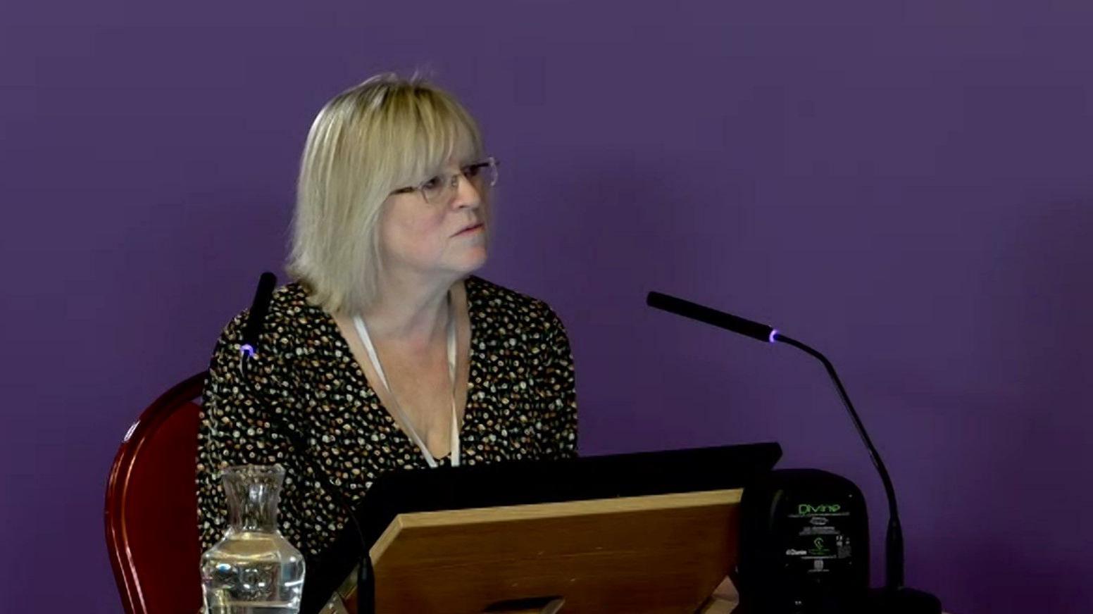 Joanne Hodson, who has shoulder length blonde hair, a fringe and glasses, sits at a witness table in front of a screen and a black microphone in front of a purple screen.