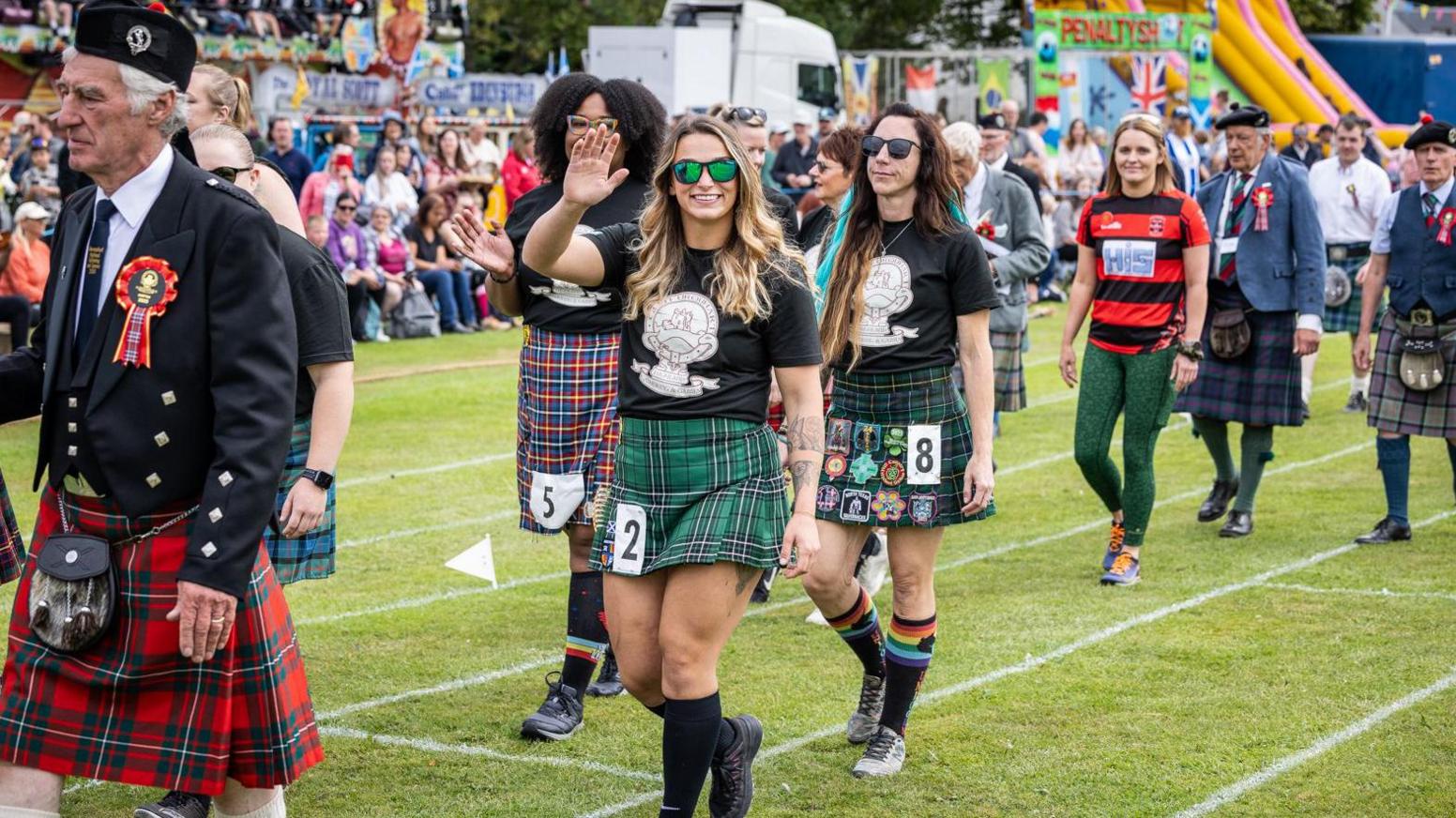 The female athletes are pictured taking part in a parade during the games. They are wearing black t-shirts and kilts of different tartans. Also in the parade are Highland games judges, wearing black jackets, kilts and hats, including Tam o' Shanters.