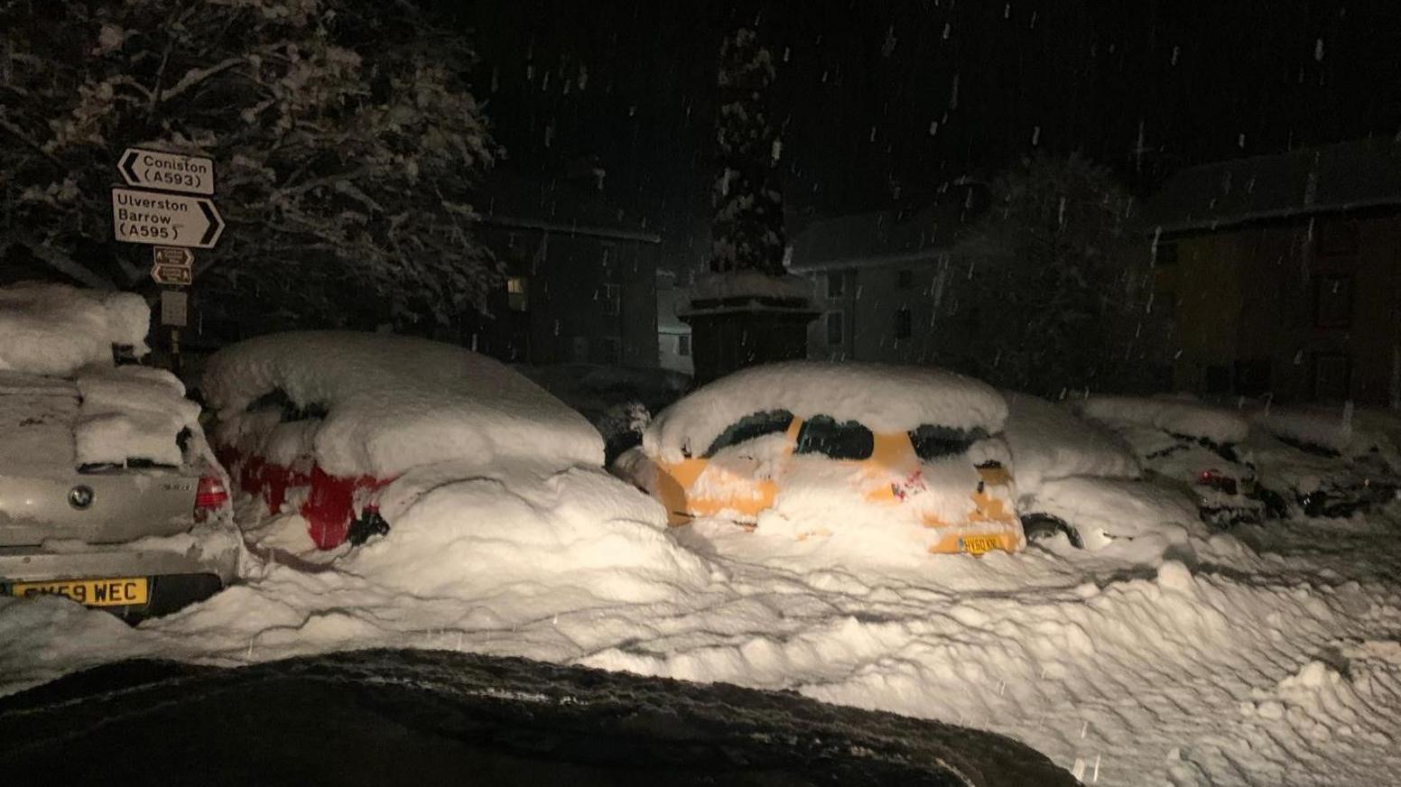 A number of cars in Cumbria have at least a foot of snow on them and are stuck deep in drifts. Behind is a roadsign pointing to Coniston in one direction, and Ulverston in the other