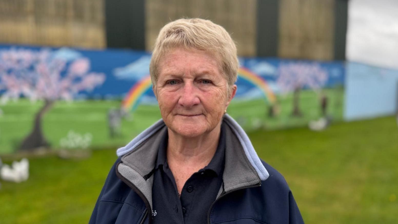 Fiona Squibb from Louth SPCA looks towards the camera with a neutral expression.  She is a woman with short, blonde hair and is wearing a black polo shirt under a navy, fleece-lined jacket. She is standing in front of a wall which is painted with a mural showing cherry blossom trees and a rainbow. 
