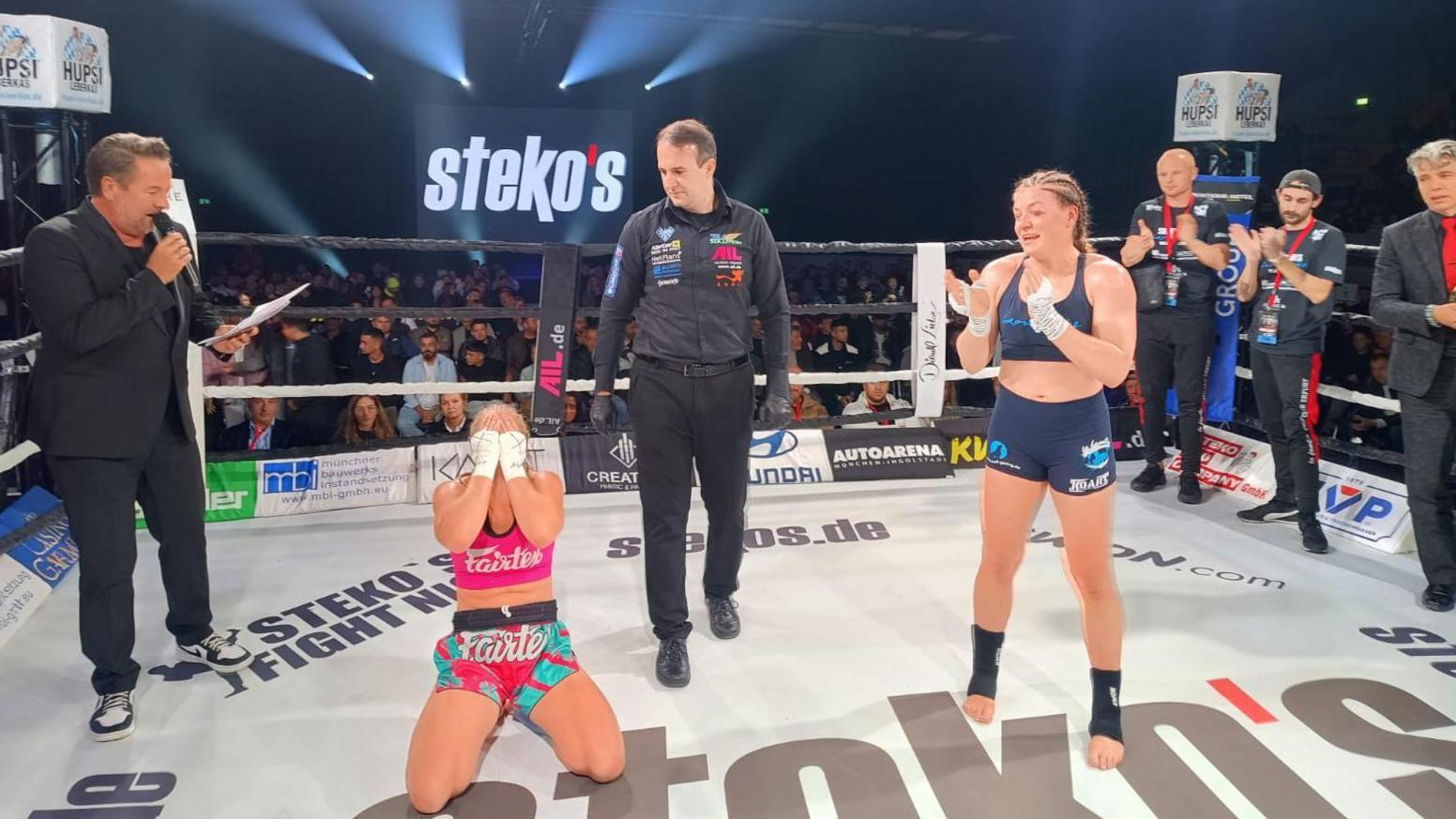 Hannah Turner on her knees covering her face with opponent Michaela Michl applauding to her right, and five black-clad men also in the boxing ring, with a watching crowd in the background