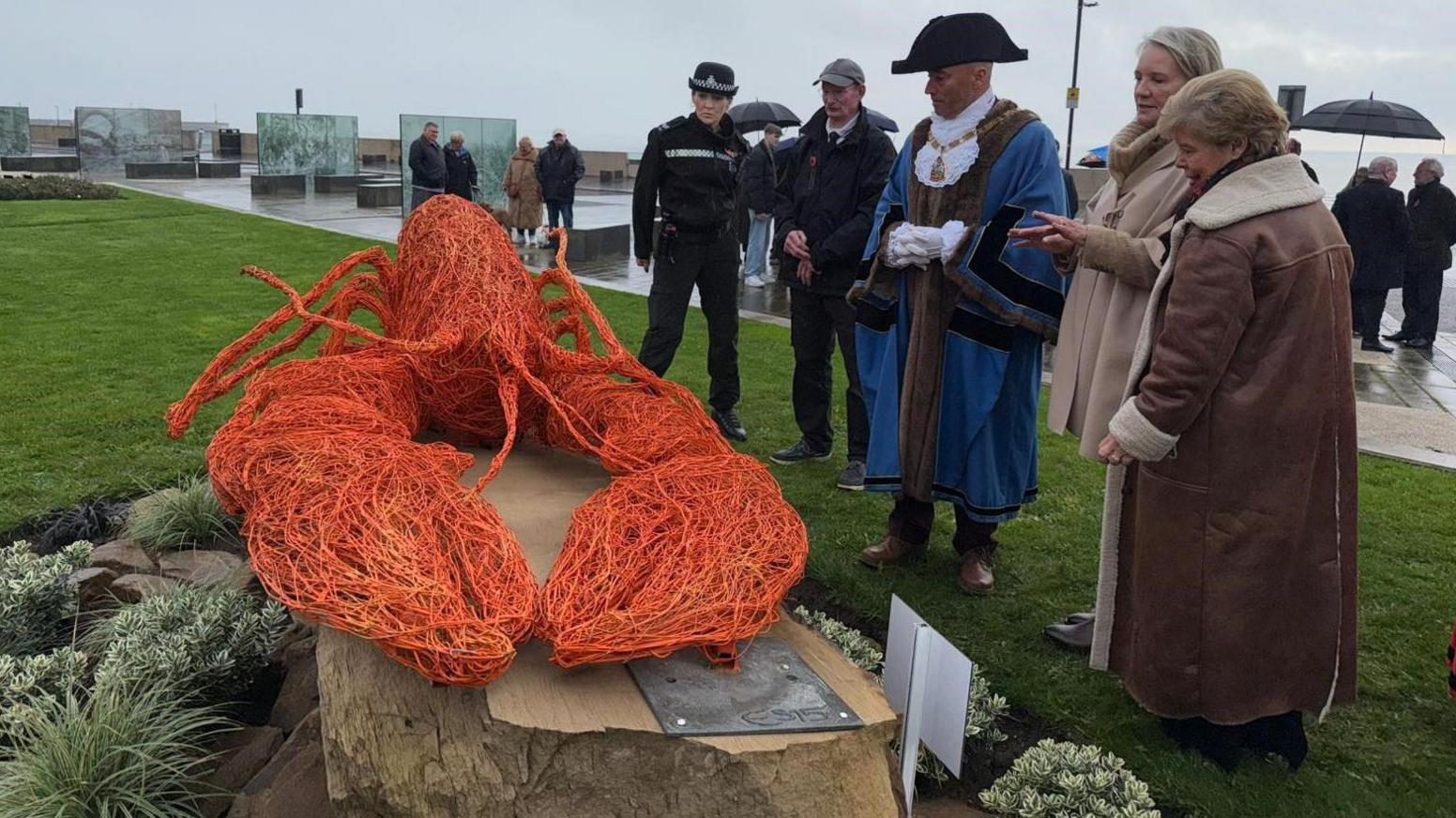 The sculpture is srrounded by a group of people who are examining it. It sits on a stone plinth surrounded by planting and the wire lobst is bright orange.