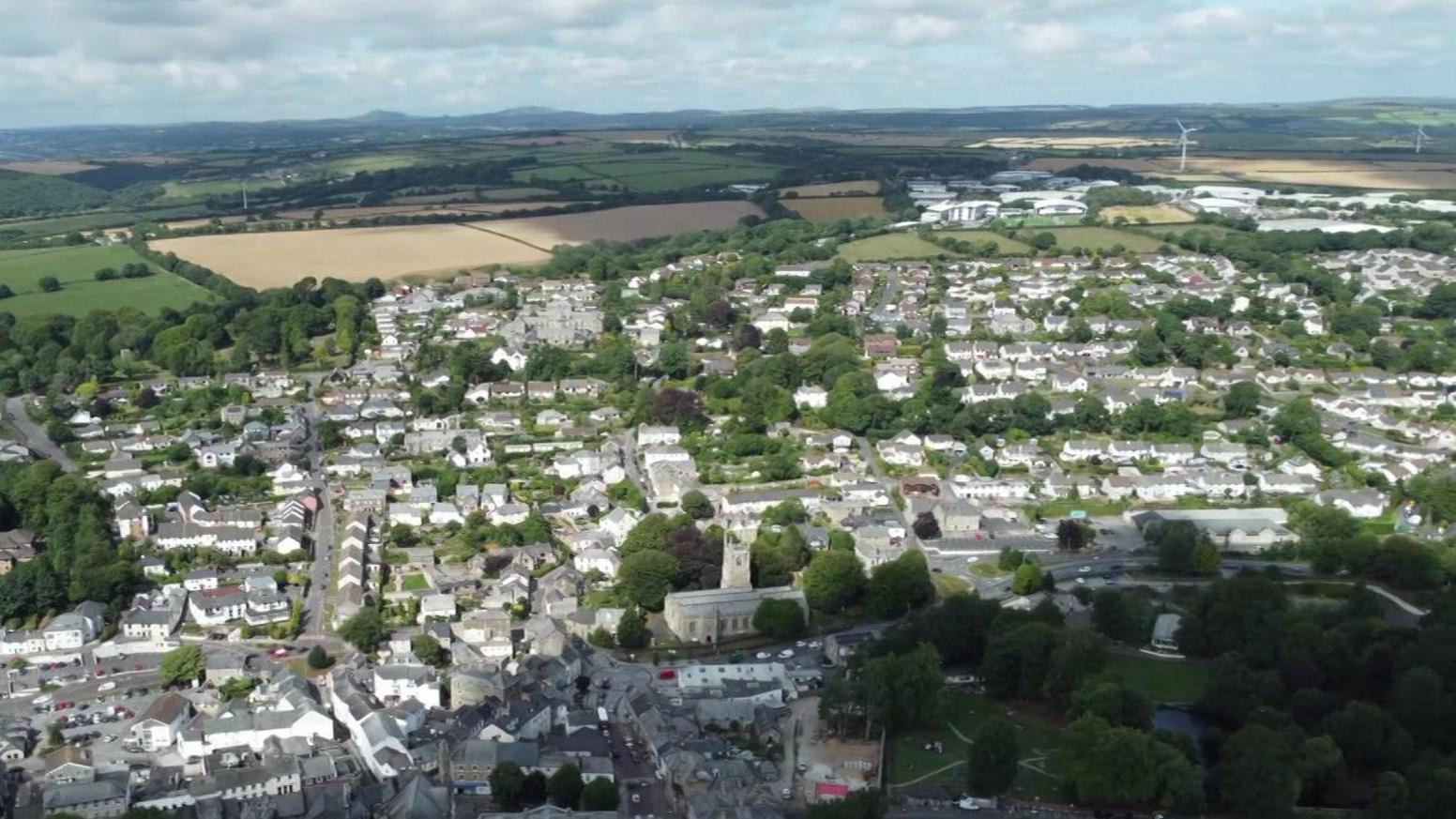An aerial drone shot of Bodmin. There are lots of houses and buildings with lots of trees and greenery around the streets and home. There are lots of fields in the background with the occasional wind turbine.