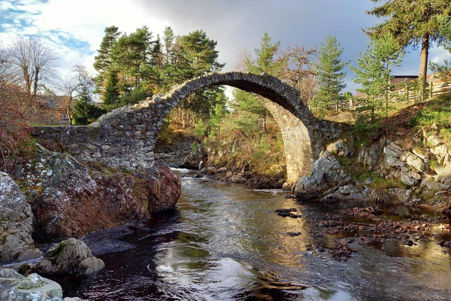 The bridge arches over a river. Sunlight hits the underside of the bridge and it crosses between two areas of rock.