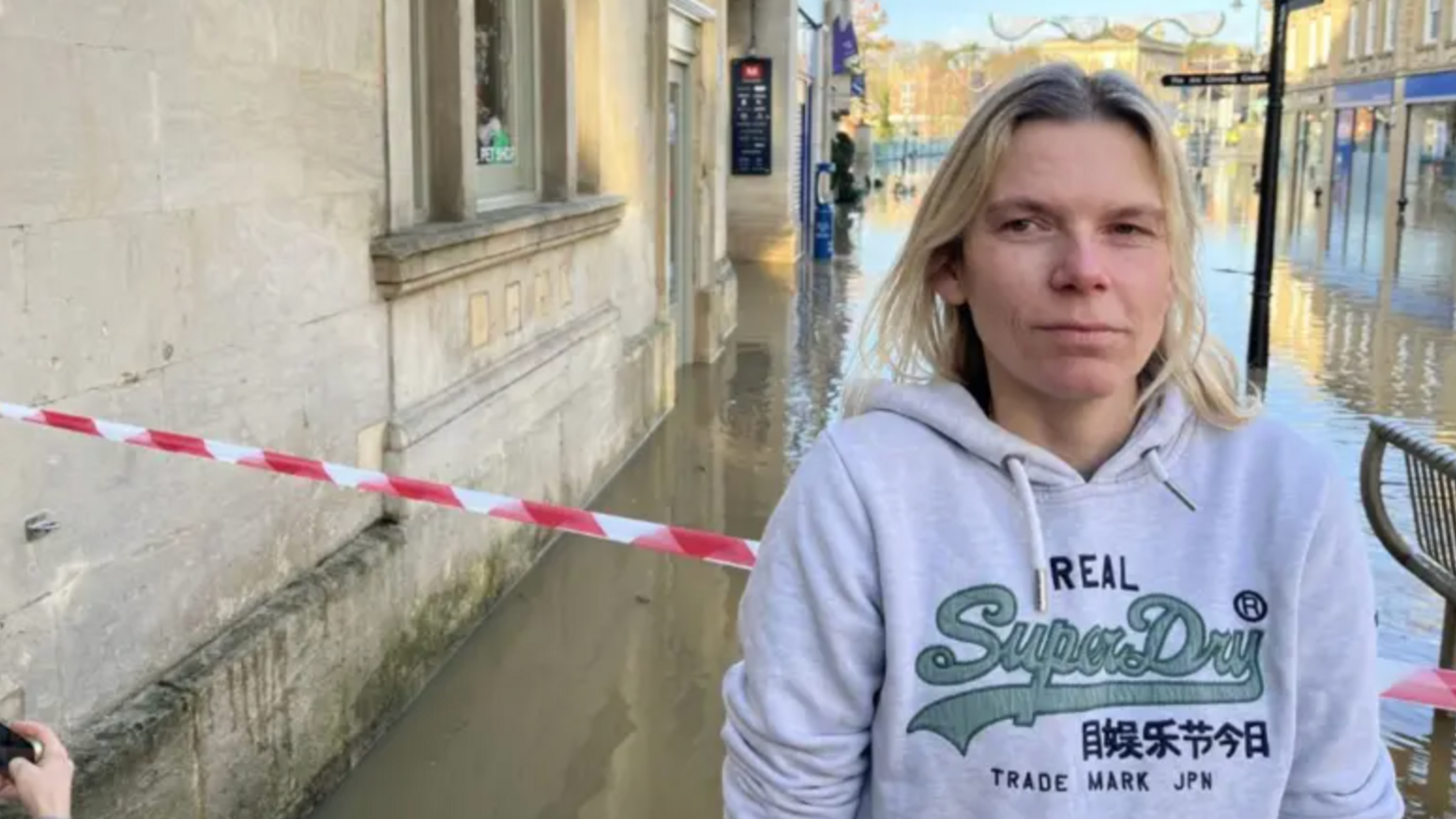 A blonde-haired woman wearing a hoodie stands in front of caution tape in front of a flooded street.