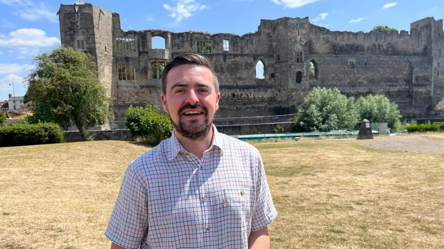 A man stands in front of Newark Castle. He is wearing a checked shirt and has dark hair and a beard. He's smiling in the sunshine.