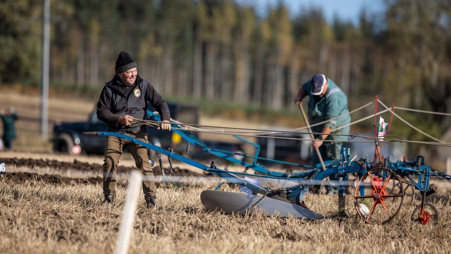 A man wearing a woolly hat works on a plough, while another man in a cap and boiler suit uses a tool to dig at the ground.