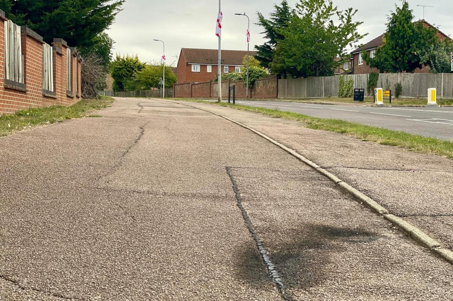 Black scorch/burn marks on a pavement. There are three lamp-posts with England flags hanging from them, and housing on the other side of wooden fencing and brick walls in the background