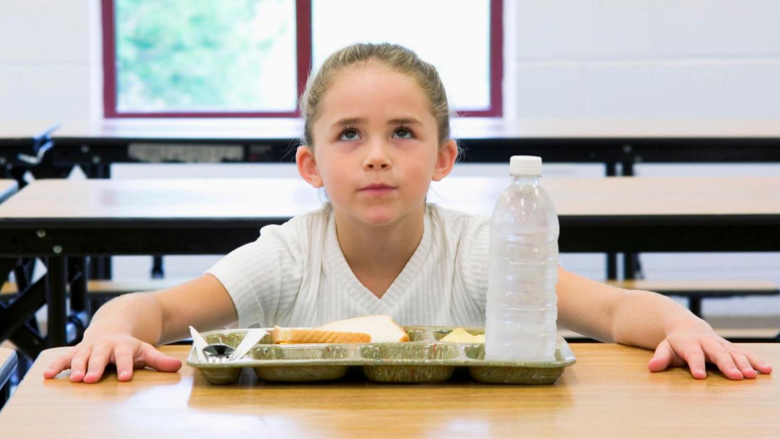 Stock image of a young girl with light hair, wearing a white top. She looks frustrated with hands on a school table in front of a tray of food and bottle of water.