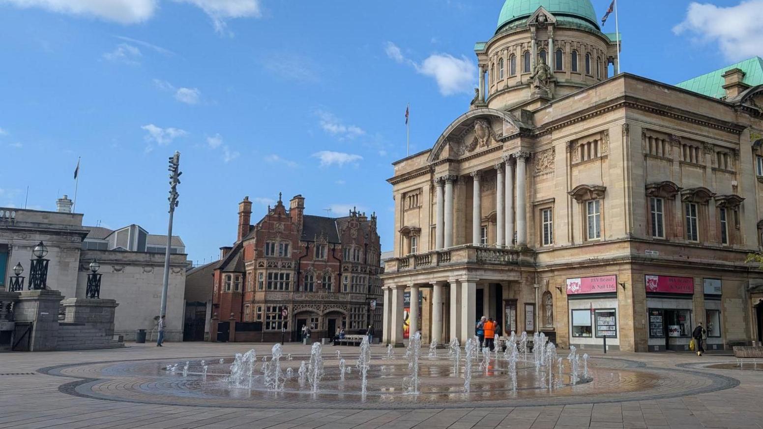 The fountain in Queen Victoria Square, Hull, is located in front of City Hall. A seating area is visible in the background.