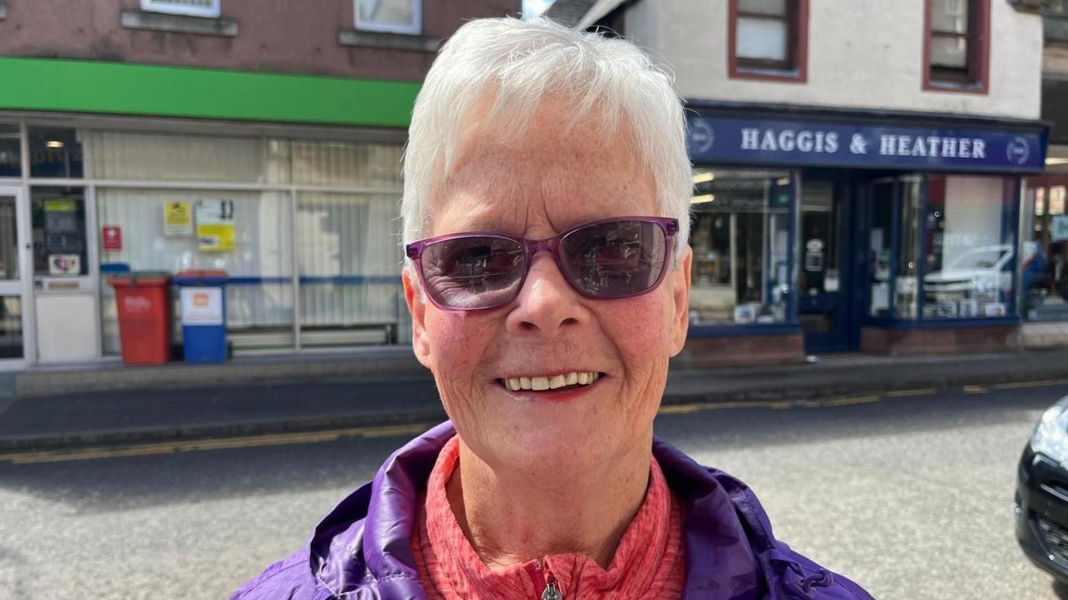 Panja is standing in Dingwall High Street. She has short white hair, is wearing purple glasses, purple jacket over a red fleece. Panja is smiling.