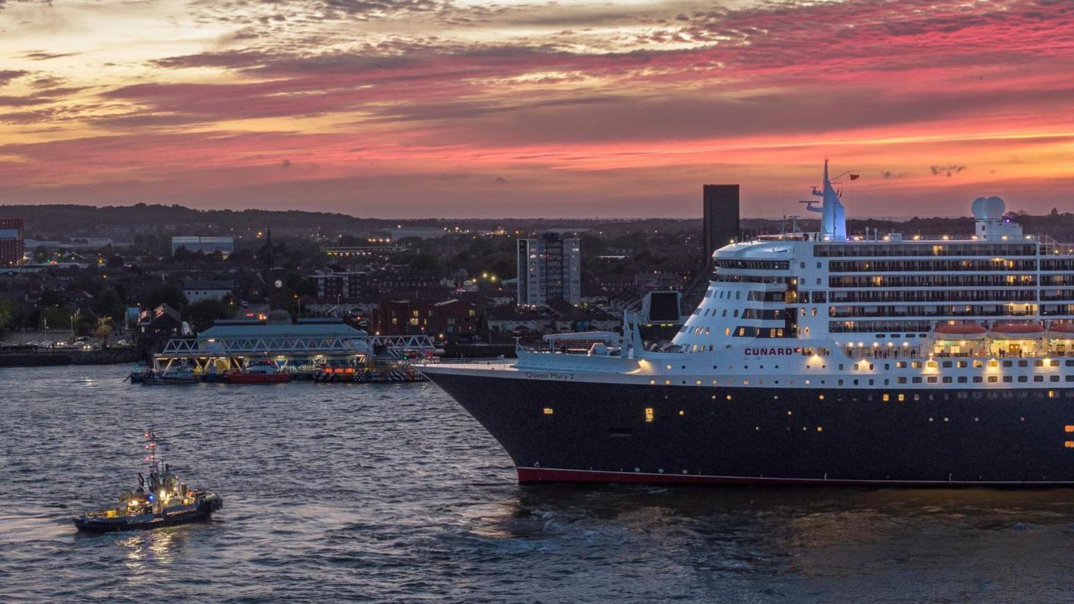The Queen Mary 2 ocean liner on the River Mersey at night. The Wirral peninsula is in the background and the sky is purple and yellow as the sun sets. A smaller boat is sailing in front.