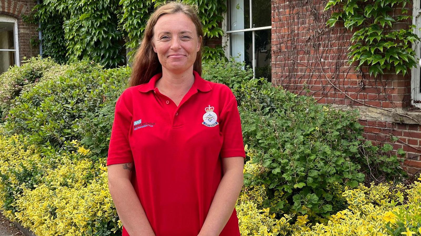 Clinical nurse specialist Vicki Bailey stands in front of some bright green shrubs. She is wearing a red polo shirt and has long ginger hair. 
