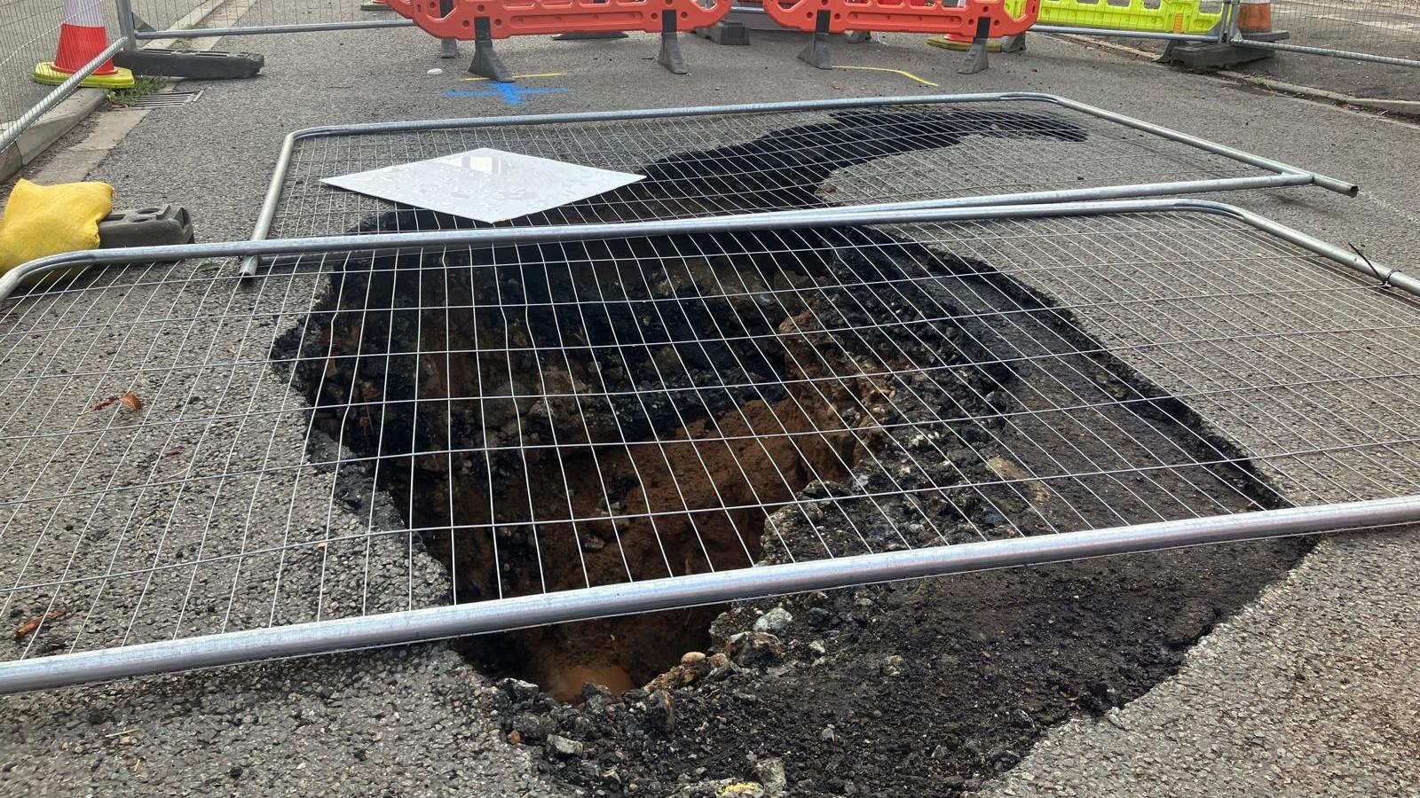 A hole in the ground covered by grey fencing, which is enclosed by cones and other barriers