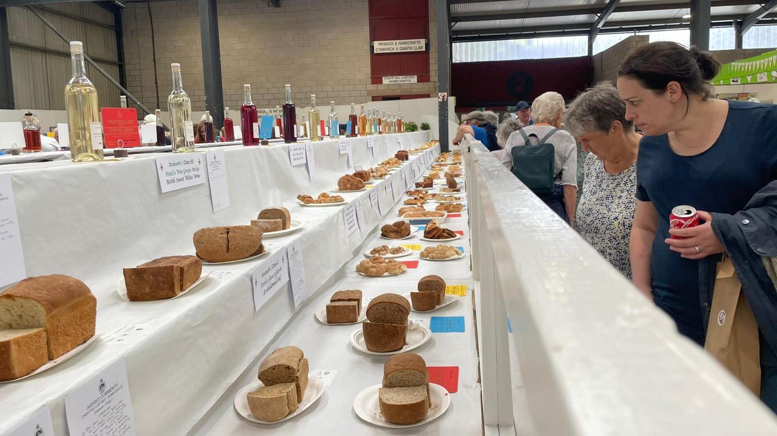 Two women looking at bread and baked goods in the horticulture tent. The woman, who is closest to the camera, has dark brown hair and is wearing a navy t-shirt. The woman behind her has short grey hair and is wearing a blue and white flower patterned blouse. 