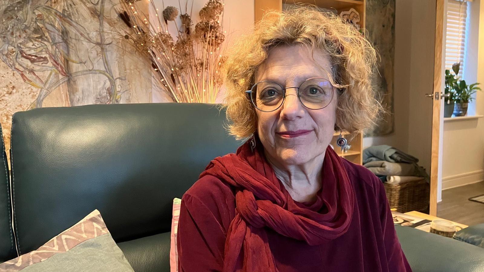 A woman wearing round glasses and red lipstick smiles at the camera. She's also wearing dangly earrings, a velvet red top and scarf. Behind her is an example of her husband's art work on the wall. 