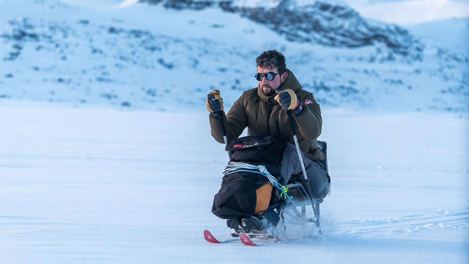 A man in an adapted sitting ski pulls himself using poles across snowy terrain. He is wearing a thick khaki jacket and sunglasses and is tied into the sit-ski with rope.
