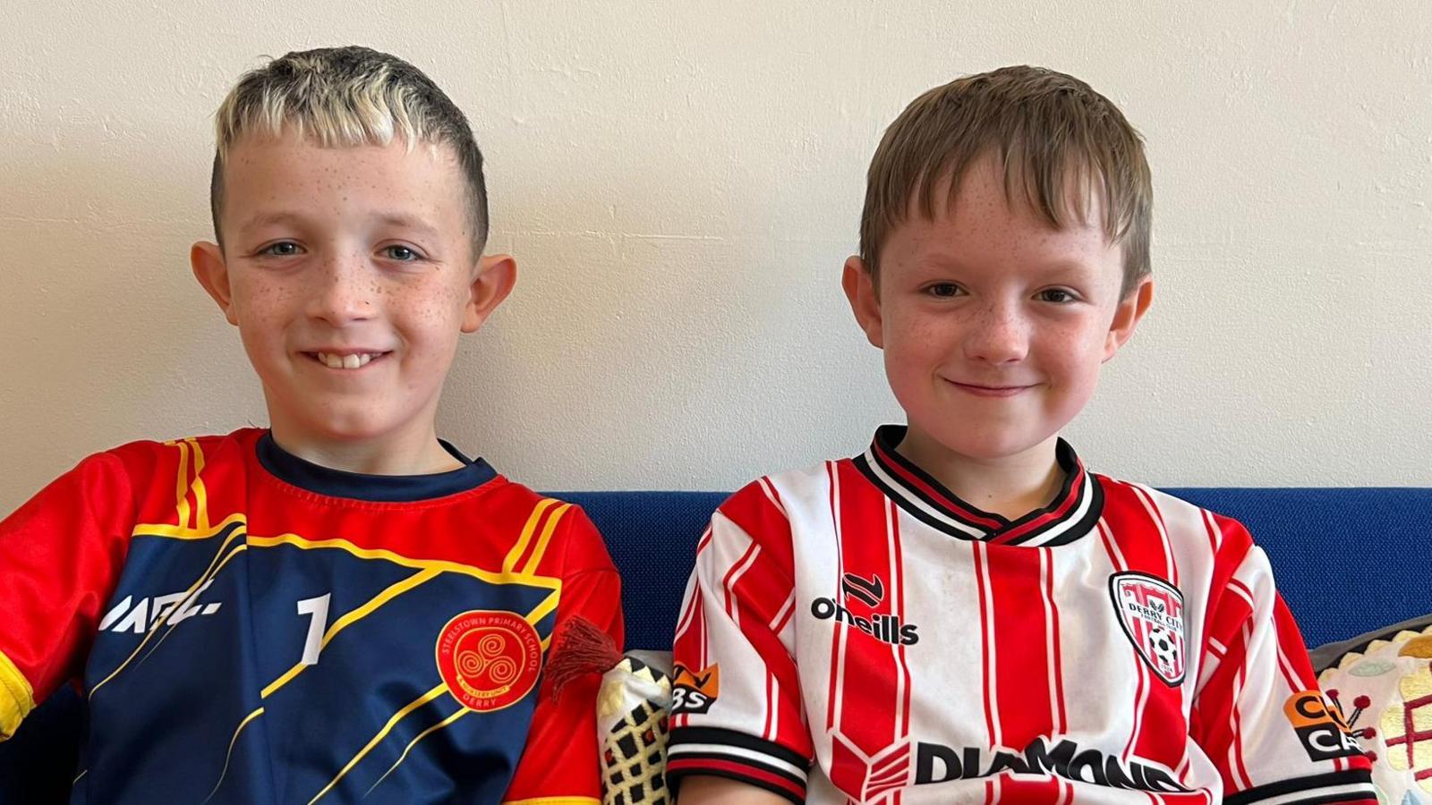 Lorcan and Charlie , wearing football tops , smile broadly. they are sitting on a blue sofa. Charlie on the right is wearing a red and white striped shirt while Lorcan on the left has a red and blue jersey with yellow streaks