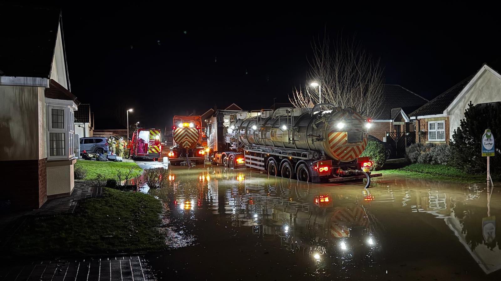 A neighbourhood with bungalows on each side of a flooded street. It is dark and emergency vehicles can be seen in the middle of the street, pumping away water.