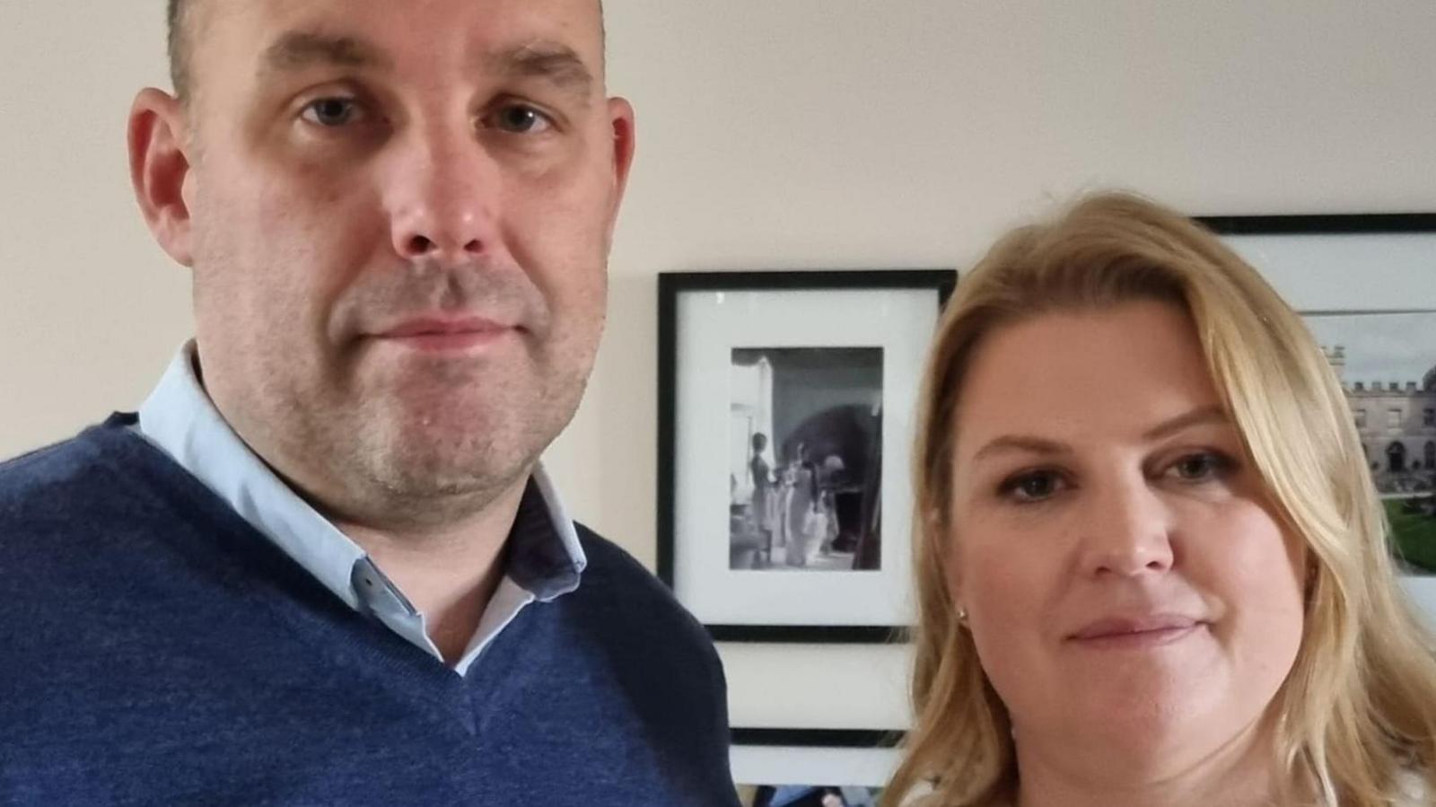 Christian and Julia Rogers are pictured in their home. Christian is wearing a navy blue jumper while Julia has a cream jacket on with buttons done up. They are standing in front of a cream wall with photographs in black frames on the wall behind them.