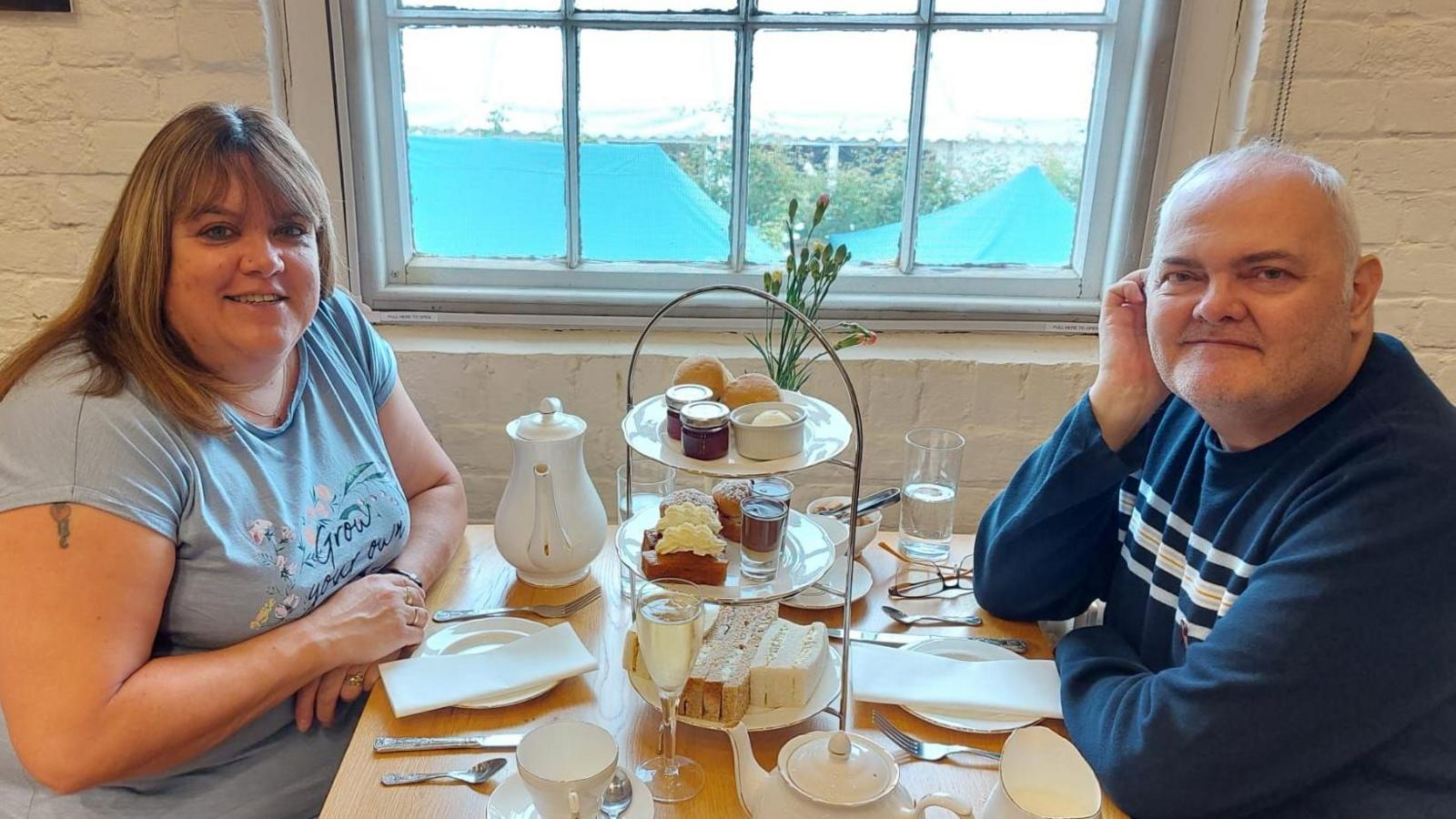 Diane (left) with long brown hair wearing a grey t-shirt and Matthew (right) with white hair and wearing a navy and white striped long-sleeved top sitting at a table by a window enjoying afternoon tea. They are smiling.