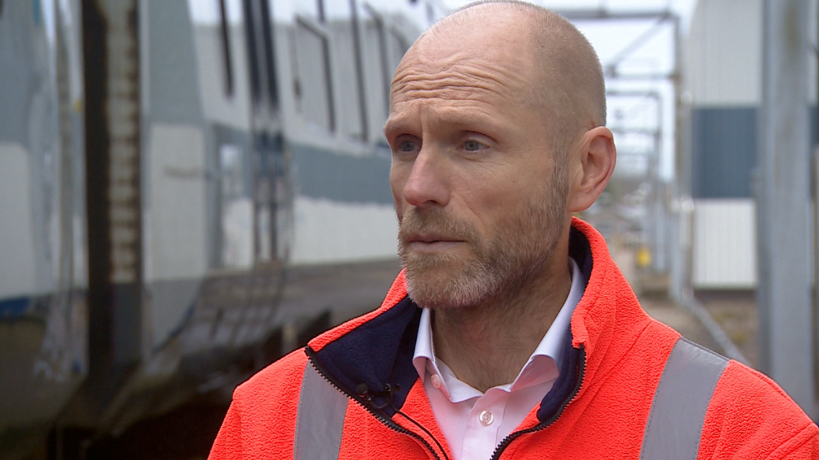 A bald man with a close cut grey beard, white shirt and orange fleecy top looks off to the left of the camera. He is standing in front of a train.