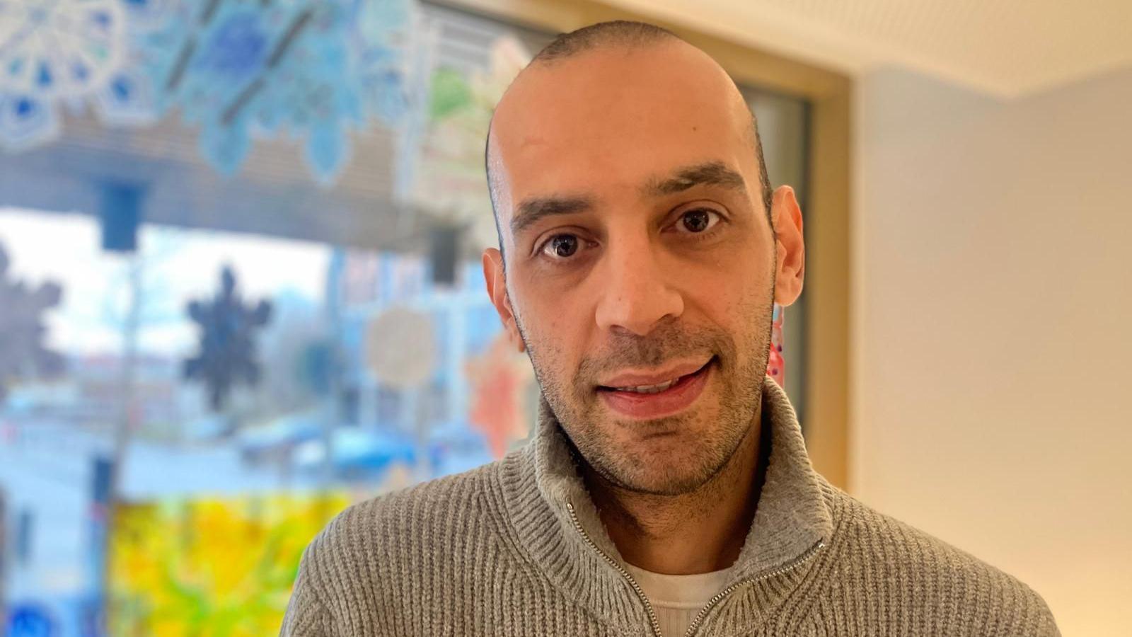 A man with a shaved head and grey jumper looks at the camera. Behind him a blue and yellow window display is blurred.